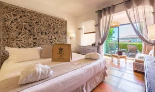 Chambre avec tête de lit en bois sculpté, peignoir posé sur le lit et baie vitrée ouverte sur la terrasse