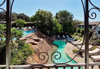 Piscine lagon bordée de transats et de pins, vue à travers une grille en fer forgé