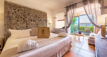 Chambre avec tête de lit en bois sculpté, peignoir posé sur le lit et baie vitrée ouverte sur la terrasse